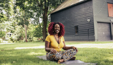 A woman sits cross-legged outside on a yoga mat. Behind her we see a large expanse of healthy-looking grass and the side of a house with a garage, which is decorated with fairy lights.
