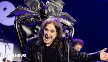 Ozzy Osbourne dressed in a black leather jacket sat on a black throne with a microphone in front of him. He has his mouth open wide as he shouts at the crowd, gesturing with his left hand. A microphone is in front of him, and a cross is hanging off his jacket.