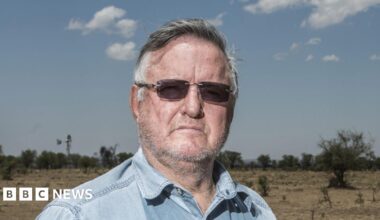 John Hume looks into the camera, wearing rimless sunglasses and a blue denim shirt