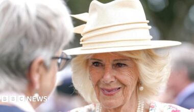 Queen Camilla wears a light coloured hat, pearl earrings and a necklace and a white dress with pink flowers.