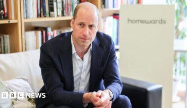 A balding man in a blue suit is sat on a sofa. He has a straight face and is clasping his hands together in front of himself. He is mostly bald with very short brown hair on either side.