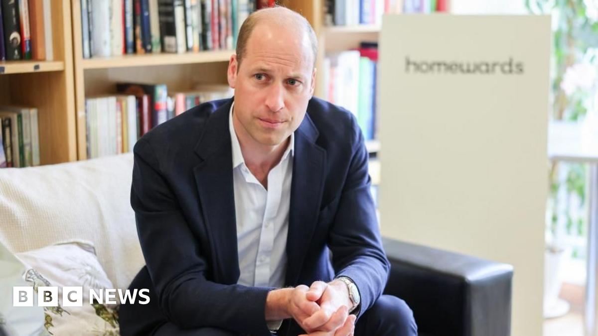 A balding man in a blue suit is sat on a sofa. He has a straight face and is clasping his hands together in front of himself. He is mostly bald with very short brown hair on either side.