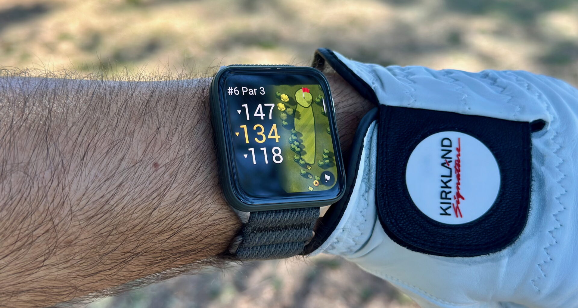 The Garmin Venu X1 on a wrist next to a golf glove, showing the yardage for the hole with fairway rough in the background.