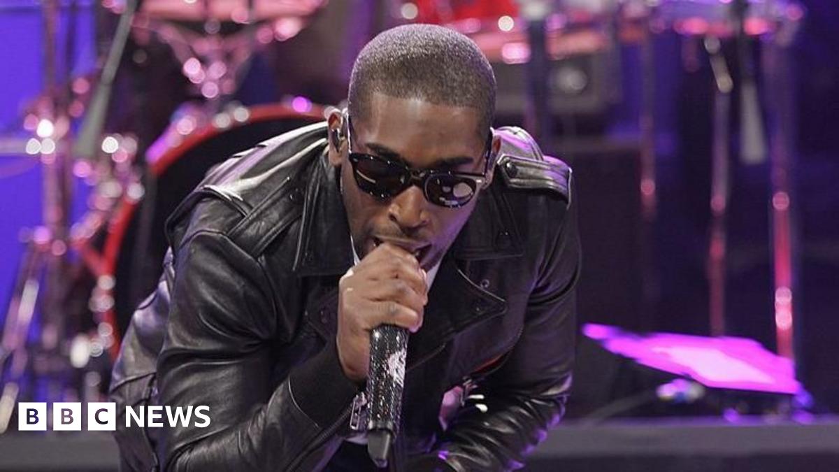 Tinie Tempah pointing left of the camera. He's smiling and wearing sunglasses and a white t-shirt while singing into a mic. Behind him is a green screen that says "Tinie Tempah - hashtag the last night out".