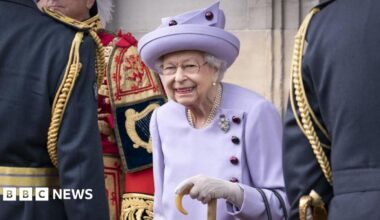 The late Queen Elizabeth wearing a lilac suit and hat , white gloves and holding a walking stick. She is wearing pearls and a broach. She is flanked by three men in royal livery but their faces are not visible.