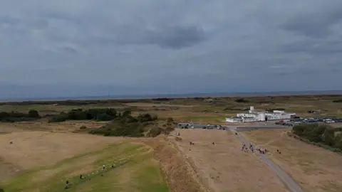 An aerial image shows common land close to Hillside Golf Club. It is a stretch of land behind a band of sand dunes leading down to the beach and the sea beyond. The white Art Deco Royal Birkdale club house is just behind the strip of common land