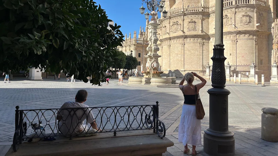 A man sits on a bench in front of a cathedral. A woman is standing taking a photo. In front of the cathedral, a sandy coloured building with a curved frontage, a decorative finished and balustrades along the balcony section. There is also a water fountain in the plaza area which has grey stone paving.