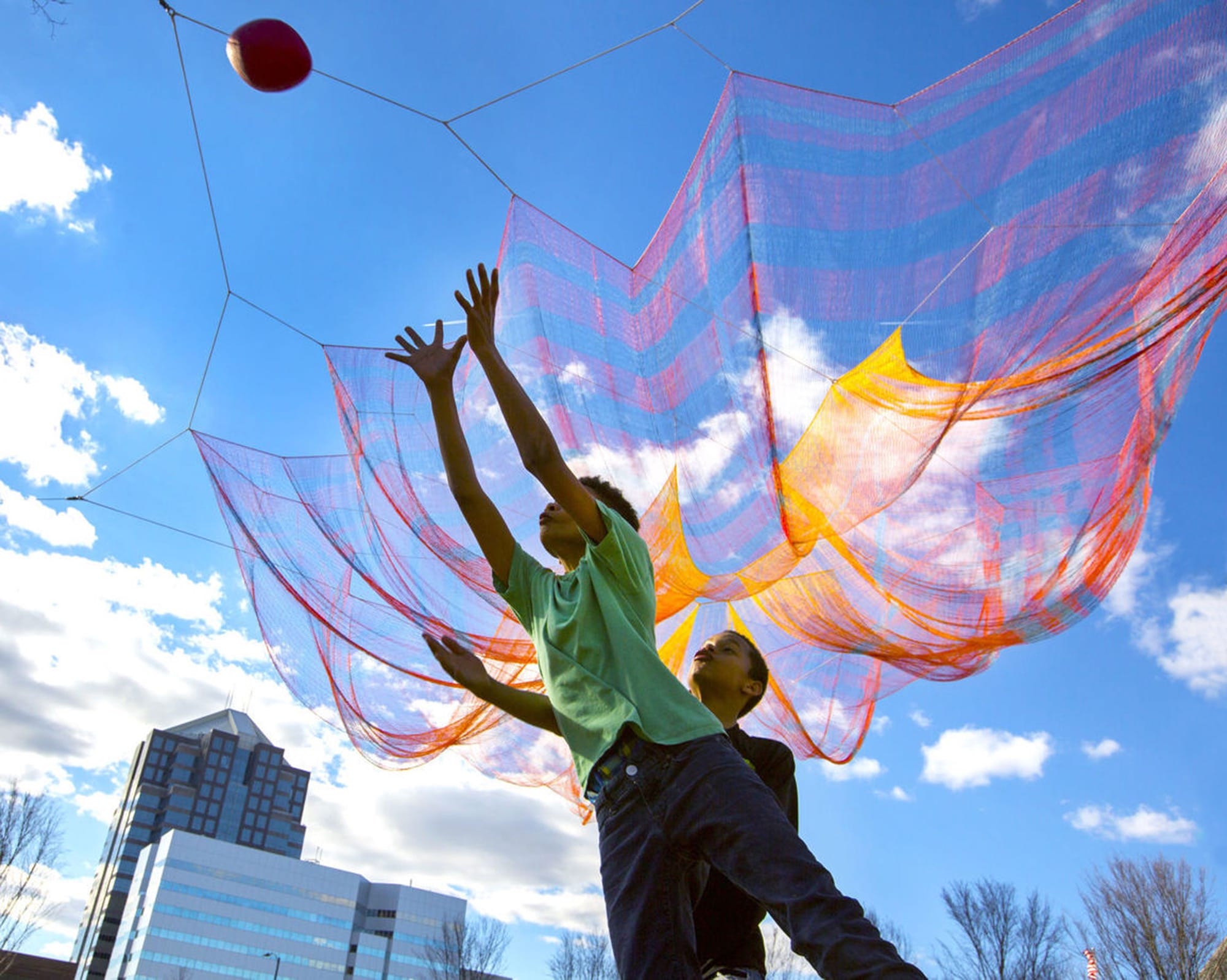 kids play basketball beneath an orange and pink Janet Echelman installation suspended in the air