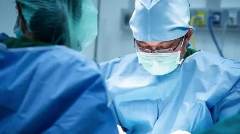 Getty Images Close up of a surgeon during an operation. He is wearing blue scrubs and a face mask. He is leaning slightly forward and looking down through a pair of black-framed spectacles. 