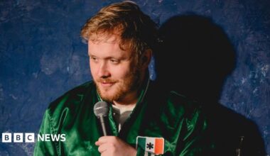 Reece Kidd on stage wearing a green jacket with an Irish flag on the left breast. He has short light brown hair and a short light brown beard. He is holding a microphone to his mouth and standing in front of a dark blue backdrop.