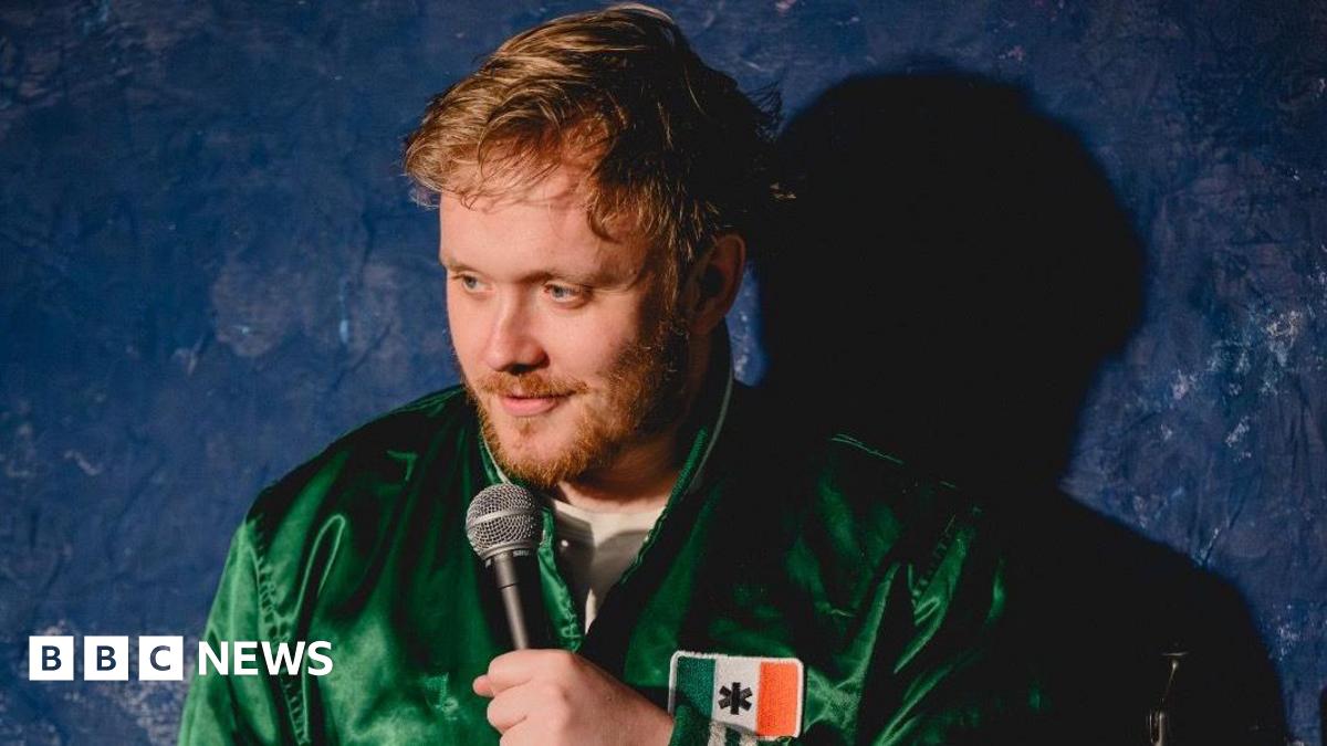 Reece Kidd on stage wearing a green jacket with an Irish flag on the left breast. He has short light brown hair and a short light brown beard. He is holding a microphone to his mouth and standing in front of a dark blue backdrop.