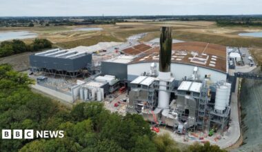 The Integrated Waste Management Facility (IWMF) at Rivenhall, which is an incinerator designed to burn Essex County Council's household waste. A drone photograph shows the domed roof of the incinerator building, covered with browned grass. There are several white hatches in the roof of the building. Next to it are several large pipes, and a 35-metre tall chimney which is covered in a reflective surface. There are green fields and trees in the distance.