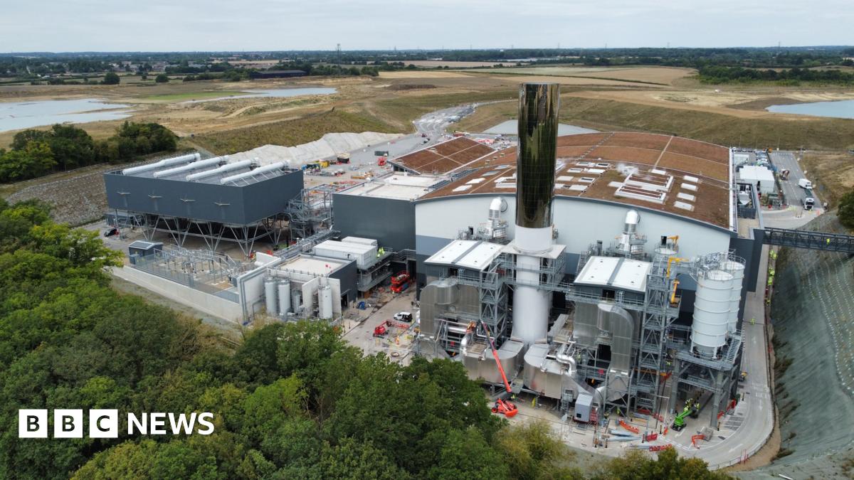 The Integrated Waste Management Facility (IWMF) at Rivenhall, which is an incinerator designed to burn Essex County Council's household waste. A drone photograph shows the domed roof of the incinerator building, covered with browned grass. There are several white hatches in the roof of the building. Next to it are several large pipes, and a 35-metre tall chimney which is covered in a reflective surface. There are green fields and trees in the distance.