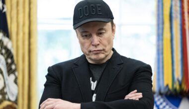 Elon Musk, wearing a baseball cap with the word DOGE, stands with while crossing his arms.