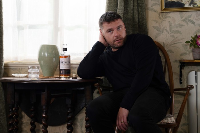 Aaron with a hand on his head as he sits with a bottle of whisky at his side in Emmerdale