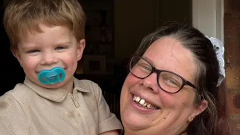 Stacey Harris holding her two-year-old son and smiling. She shows her teeth and wears black-rimmed glasses leaning her head against the side of a door. Her son has a turquoise dummy in his mouth and wears a beige top.