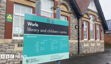 A blue and black sign outside Worle Library. The building is made of bricks with tarmac around it.