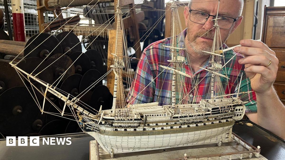 A man cleans a model ship made of ivory using a tiny paint brush