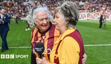 Opera singer Lesley Garratt holds a BBC Radio Leeds branded microphone and has an emotional facial expression. A female member in a Bradford City football top of the Bantam of the Opera choir smiles and talks to Lesley as they stand on the football pitch.