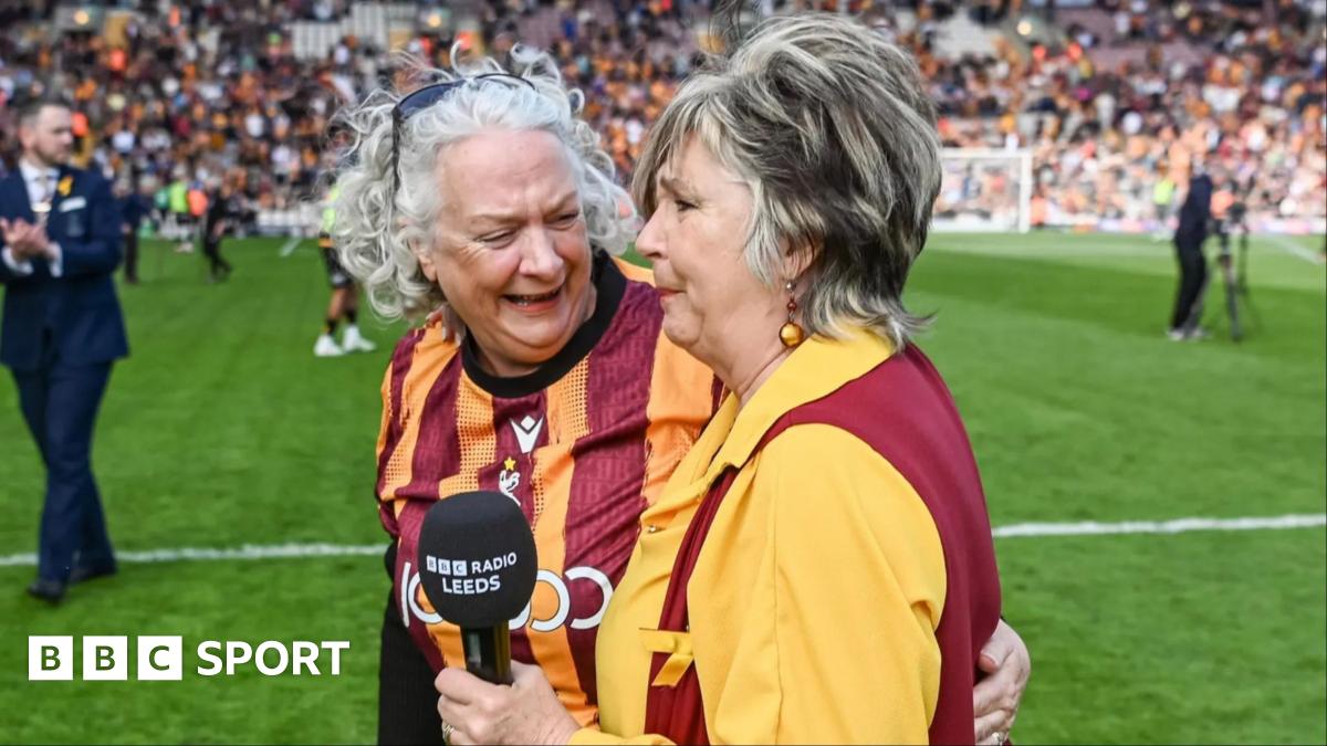 Opera singer Lesley Garratt holds a BBC Radio Leeds branded microphone and has an emotional facial expression. A female member in a Bradford City football top of the Bantam of the Opera choir smiles and talks to Lesley as they stand on the football pitch.