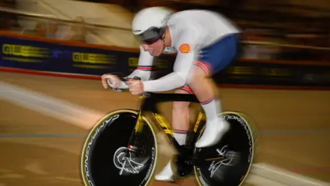 UEC A young man in white and blue GB track cycling kit and white helmet racing on his black bike in a velodrome. The background is blurred with some spectators watching. 
