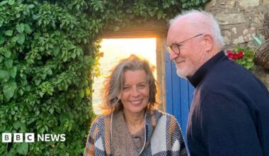 Sue Butler, with blue eyes and grey hair and a loose white, blue and orange poncho style top, with her husband Eddie Butler, a tall man with grey hair and glasses and a navy blue turtle neck sweater. They're stood in front of a stone house with an open blue door and a wall of green leaves