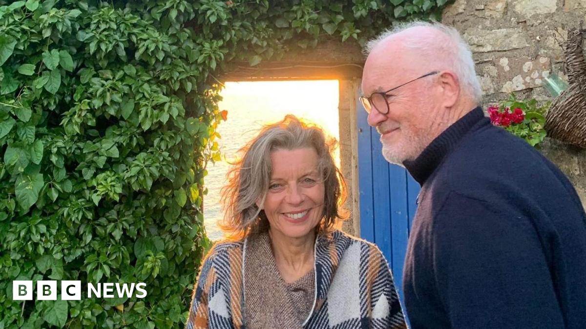 Sue Butler, with blue eyes and grey hair and a loose white, blue and orange poncho style top, with her husband Eddie Butler, a tall man with grey hair and glasses and a navy blue turtle neck sweater. They're stood in front of a stone house with an open blue door and a wall of green leaves
