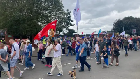 A group of people waving flags leave the side of Ormeau Park, on the Ormeau Embankment.
