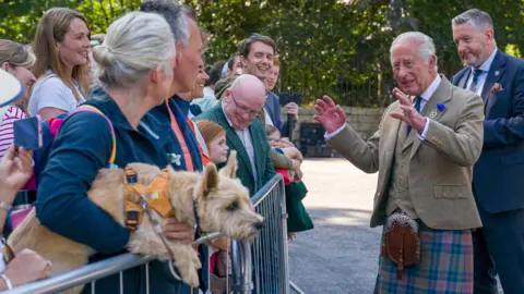 PA Media King Charles III meets members of the public after carrying out an inspection of the Balaklava Company, 5th Battalion, The Royal Regiment of Scotland, at the gates of Balmoral. He is smiling and holding his hands in front of him while talking to members of the public who are standing behind a metal barrier.