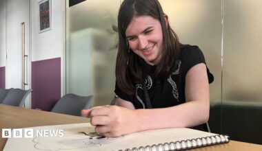 Lila in an office, smiling with her head down focused on drawing a cartoon character. She is smiling and has long black hair and is wearing a dark top.