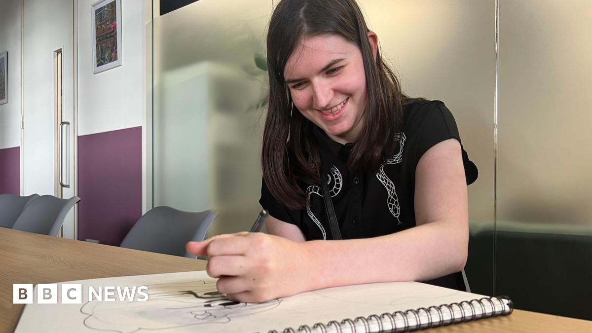 Lila in an office, smiling with her head down focused on drawing a cartoon character. She is smiling and has long black hair and is wearing a dark top.