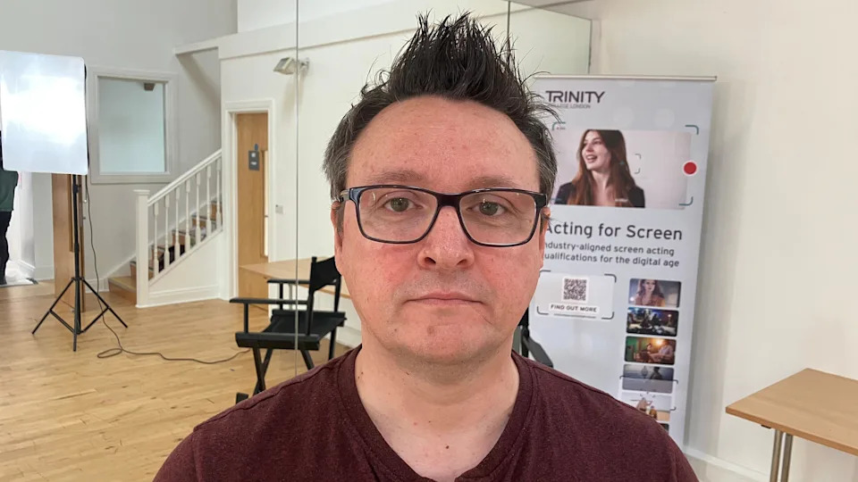 A man with spiky brown hair, wearing a burgundy t-shirt and glasses. 