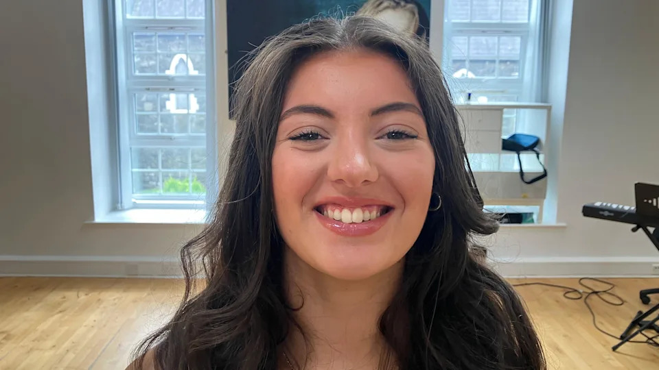 A young woman with long brown hair, smiling at the camera. She is standing in a room with a white wall and two long windows in the background. 