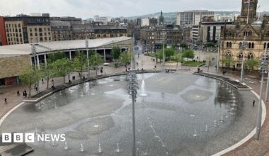 A large public square featuring a circular water feature, with multiple jets spraying water at varying heights. The water gathers to create a mirror effect. Tall light poles surround the fountain, with trees and benches lining the perimeter. People can be seen walking and sitting around the edges of the fountain. In the background, a wider city centre can be seen with a mix of modern and older buildings.