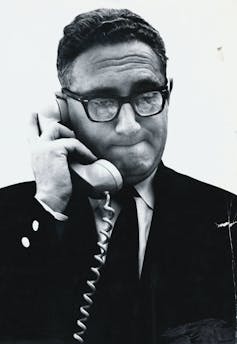 A man in glasses holds a phone next to his ear.