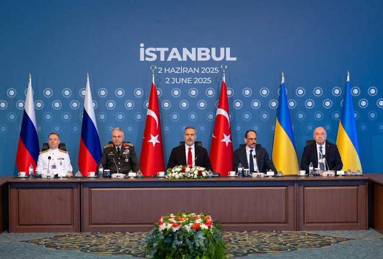A group of men sit at a desk behind which various flags are seen.