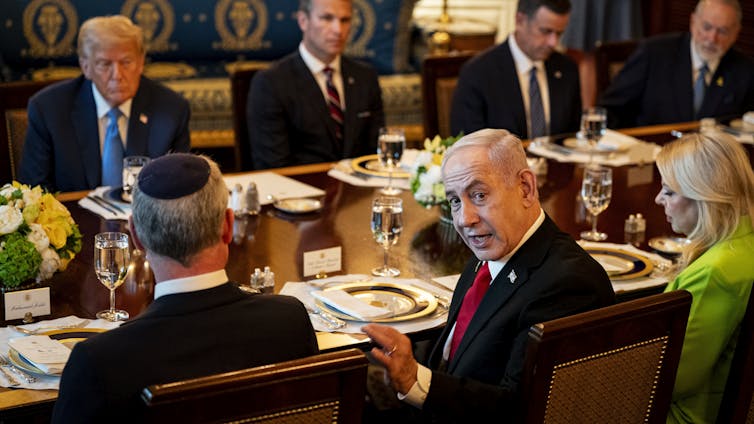 Benjamin Netanyahu turns and looks at the camera while sitting at a table in the White House with US president Donald Trump and others.