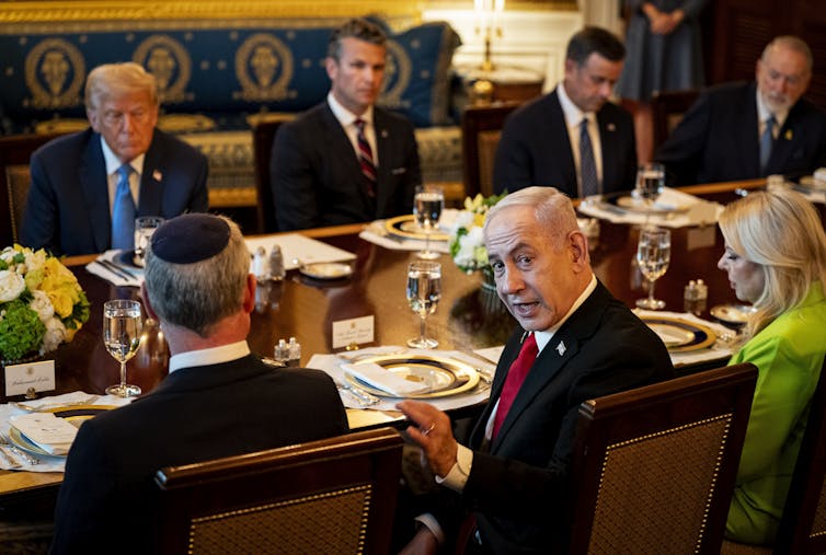 Benjamin Netanyahu turns and looks at the camera while sitting at a table in the White House with US president Donald Trump and others.
