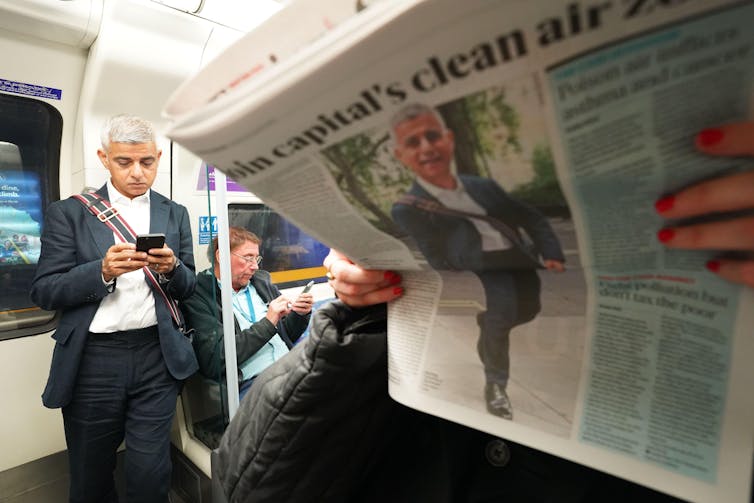 Sadiq Khan on the tube while someone reads a newspaper with his picture on one of the pages.