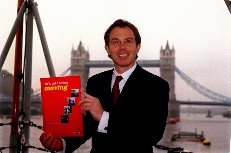 Tony Blair standing in front of Tower Bridge in London holding a document titled 'Let's get London moving'.