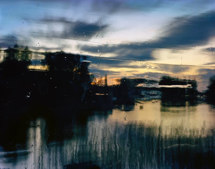 A blurry, impressionistic photograph showing a post-sunset sky streaked with clouds above a lake and trees.