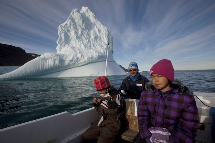 A man and two children sit in a boat with a large iceberg behind them.