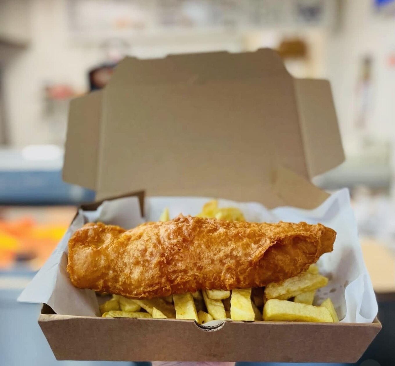 A golden piece of battered, deep fried fish sits on top of a portion of chunky, chip shop style-chips. The fish and chip shop is blurry in the background.