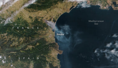 A satellite image shows a wide view of France and Spain, with the blue Mediterranean Sea to the right. A plume of gray-white smoke billows southeast from France and over the sea.