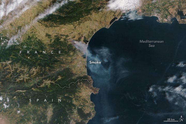 A satellite image shows a wide view of France and Spain, with the blue Mediterranean Sea to the right. A plume of gray-white smoke billows southeast from France and over the sea.