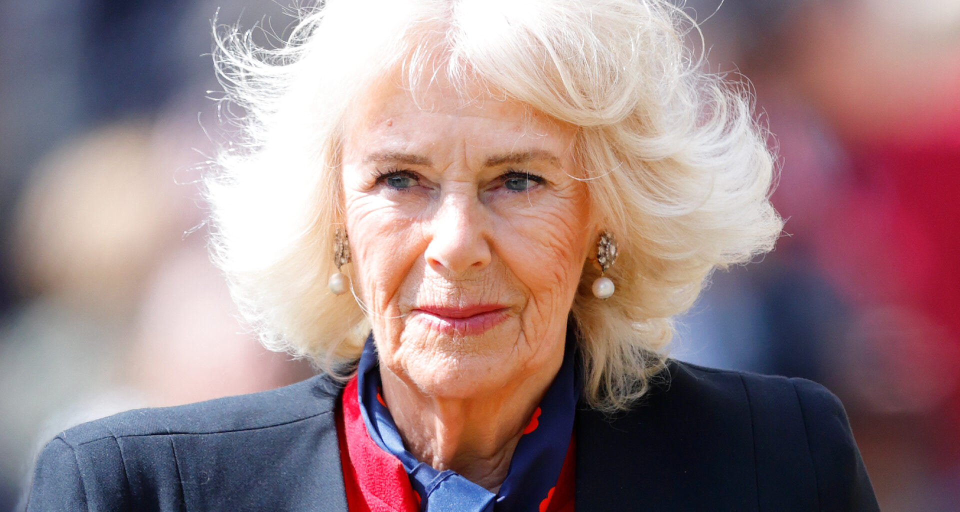 Queen Camilla wears a black blazer and a red dress and has blonde hair which is in an upheaval in the wind