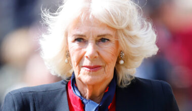 Queen Camilla wears a black blazer and a red dress and has blonde hair which is in an upheaval in the wind