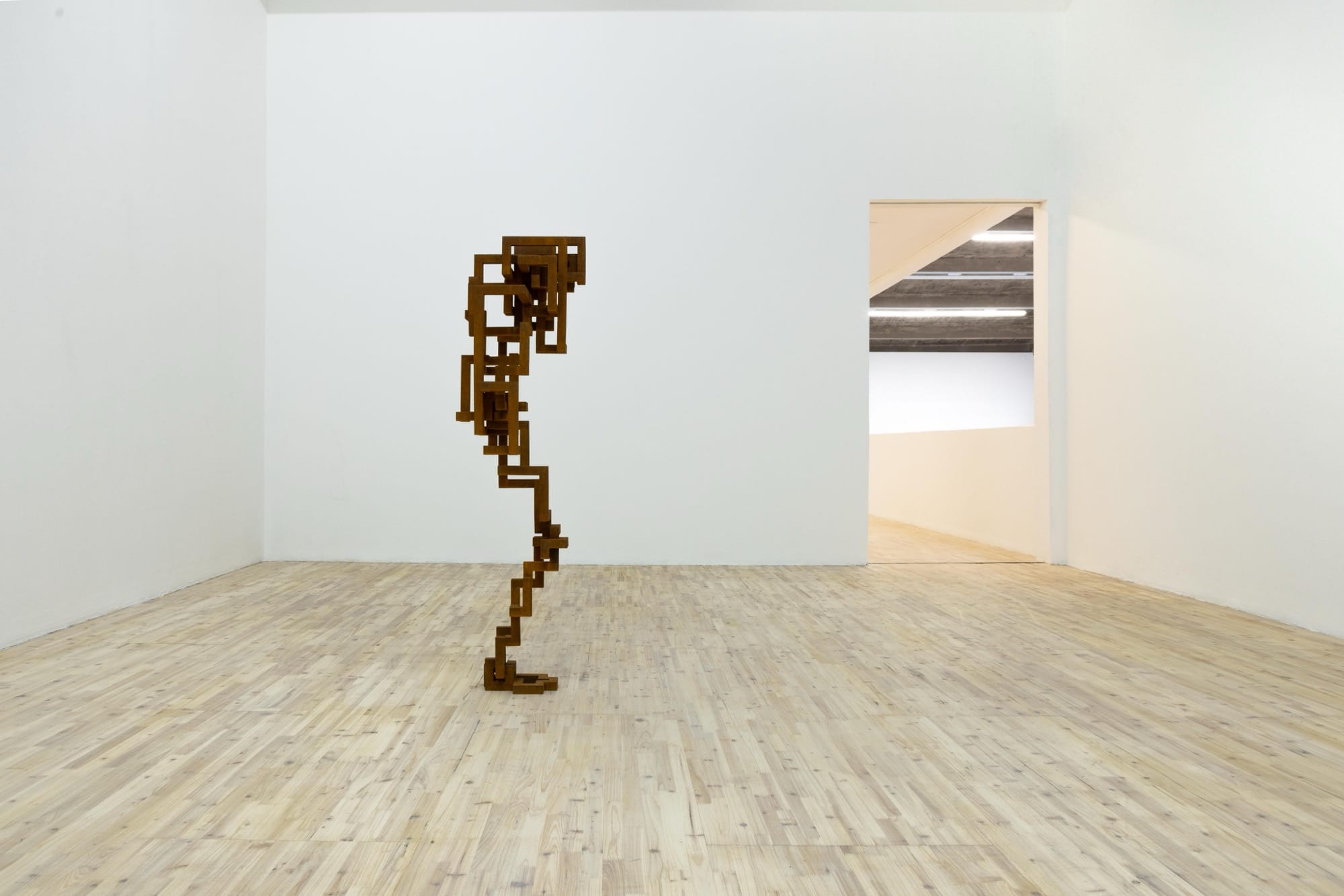 a sculpture by Antony Gormley installed in a white gallery space, depicting a blocky, abstract figure resembling a human with their head in their hands