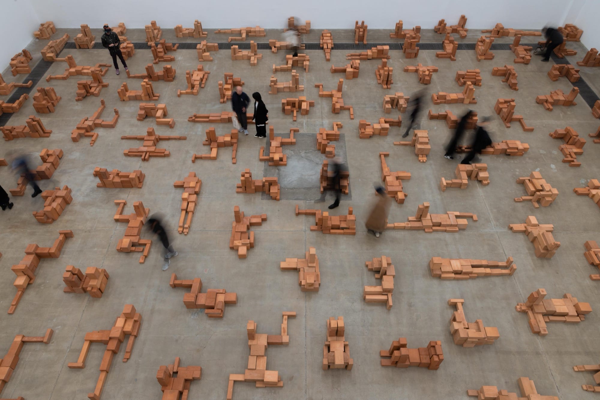people walk through a room-size art installation by Antony Gormley of blocky terracotta forms resembling human figures in various positions