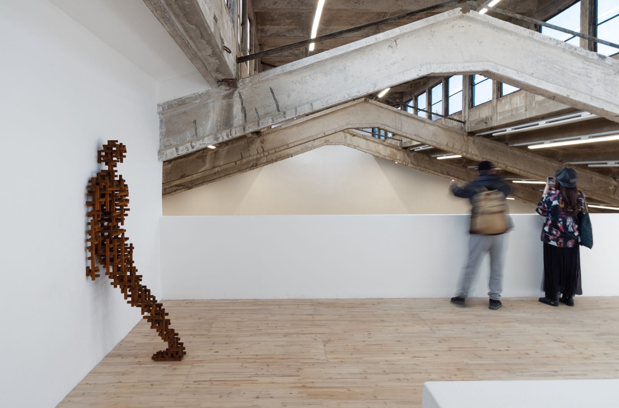 a gallery with two visitors standing near an overlook, with a sculpture by Antony Gormley installed on the wall to the left, of a blocky abstract figure resembling a human leaning face-forward on the wall